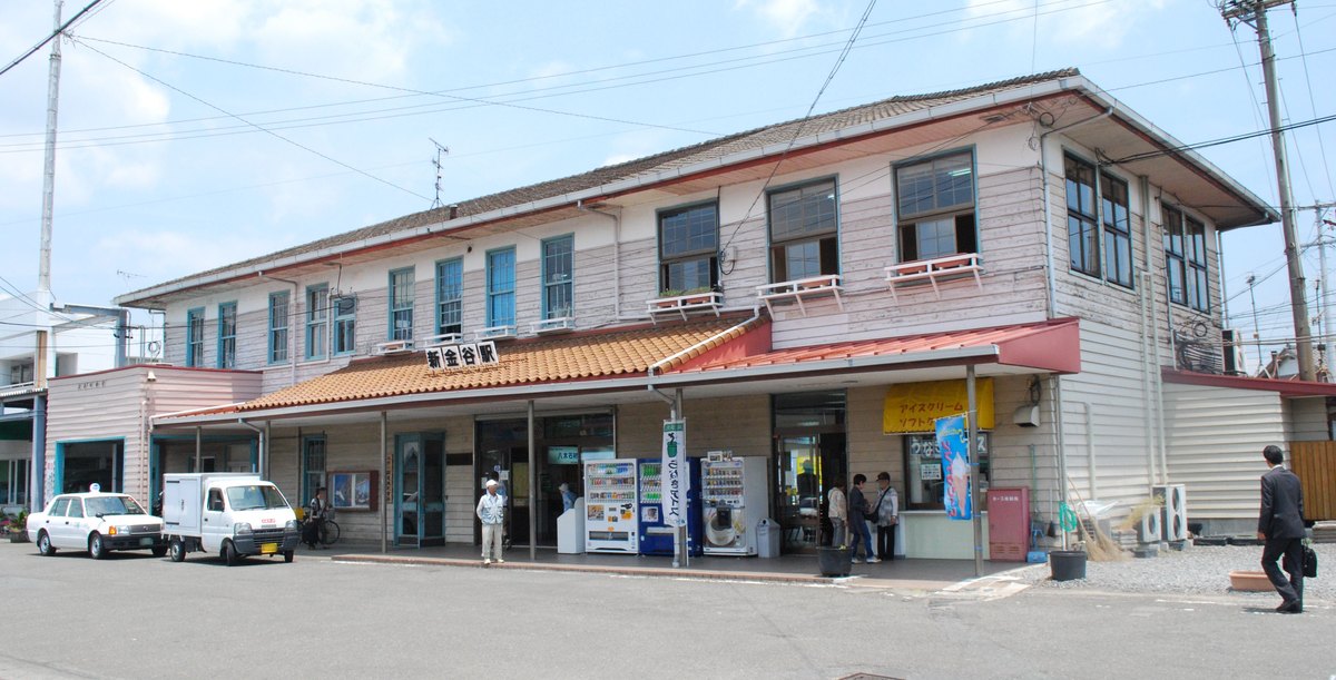新金谷站的兩層樓木造站舍：粉白色橫板外牆、橘色瓦片的門廊屋簷上掛著「新金谷駅」站名牌，門口有自動販賣機，站前停著計程車