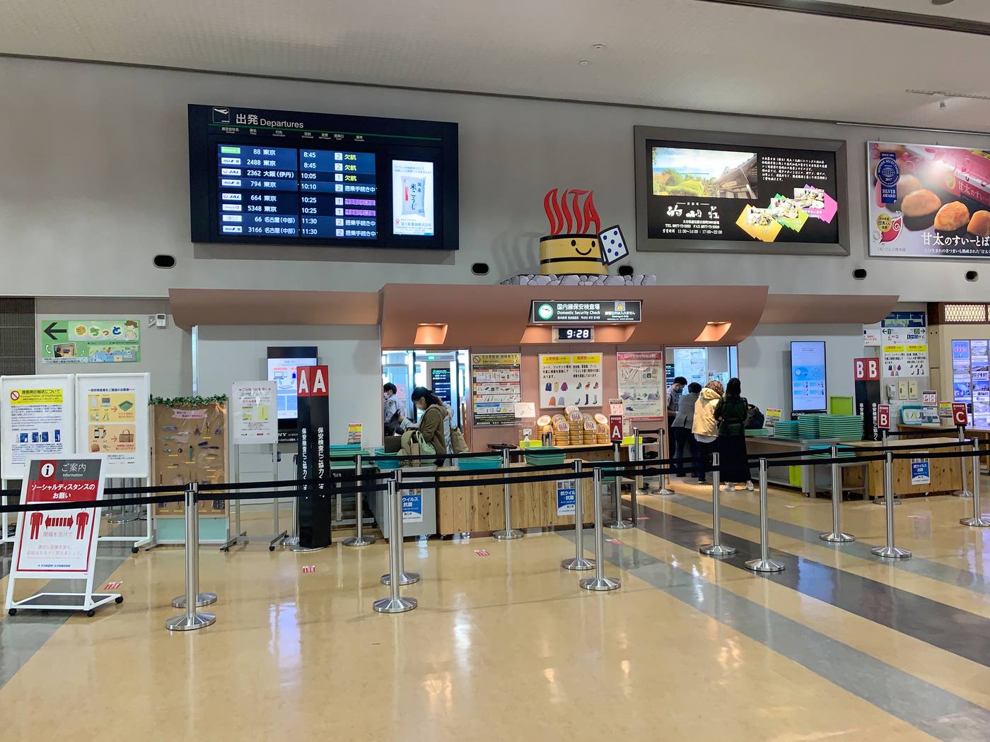 The departure floor at Oita Airport. A dark green sign above the screening lanes reads "Domestic Security Check" in Japanese and English, with a digital clock below it showing 9:28 and lane markers A, B and C to either side. Three notice boards are mounted on the wall, the middle one showing colour pictograms of coats and boots, and stacks of round wooden-tub-shaped trays printed OITA sit in front of the screening tables.