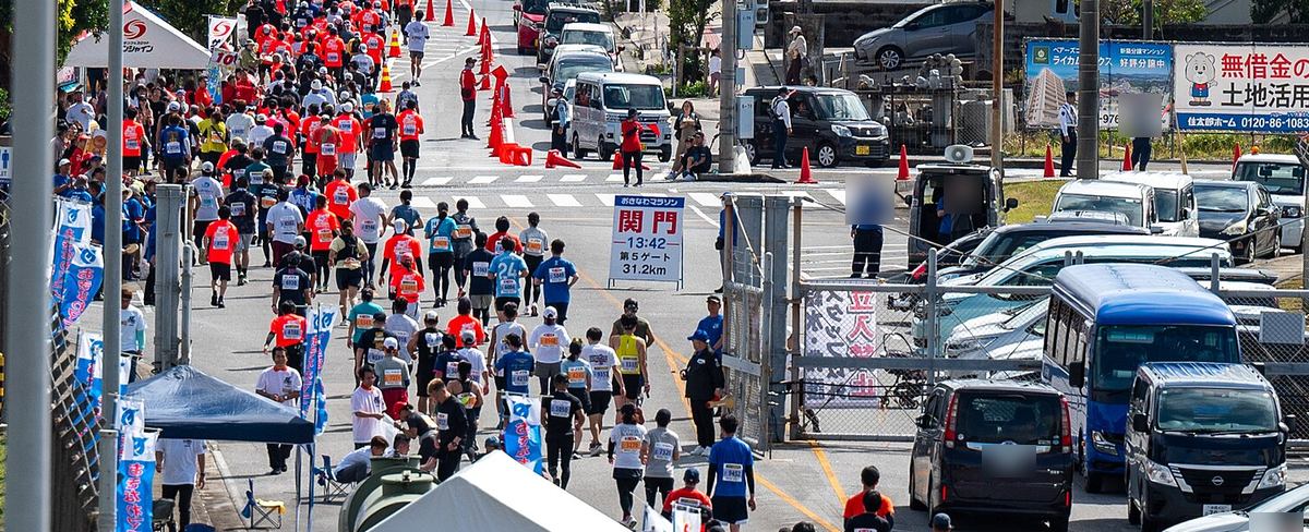 嘉手納基地第 5 門外的道路,大批跑者背對鏡頭往前跑,路中央立著白底看板,紅字寫「関門」,下面是「13:42 第5ゲート 31.2km」,左邊是補給站的藍色帳篷與旗幟,右邊是圍欄後停放的車輛