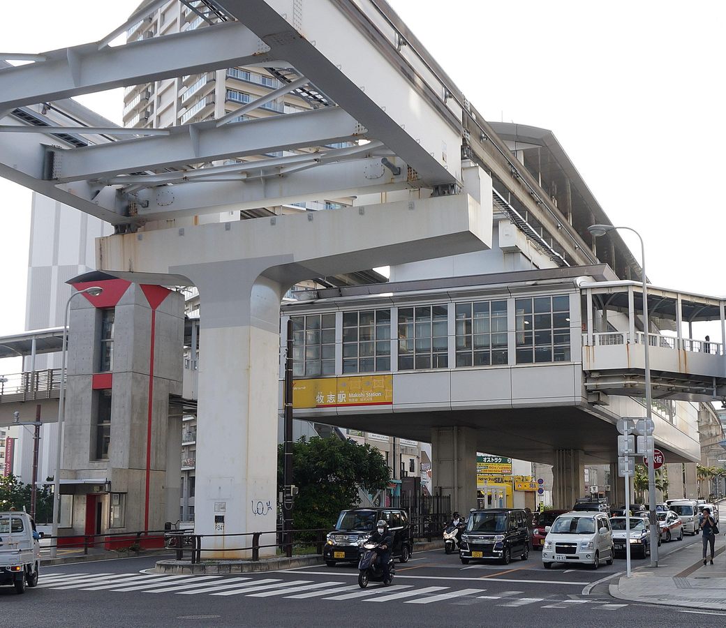 單軌電車牧志站:高架軌道與車站橫跨路口,車站外牆黃色站名板寫「牧志駅」,路口停著等紅燈的車輛與機車,右側是架在路口上方、有屋頂和欄杆的高架人行天橋平台