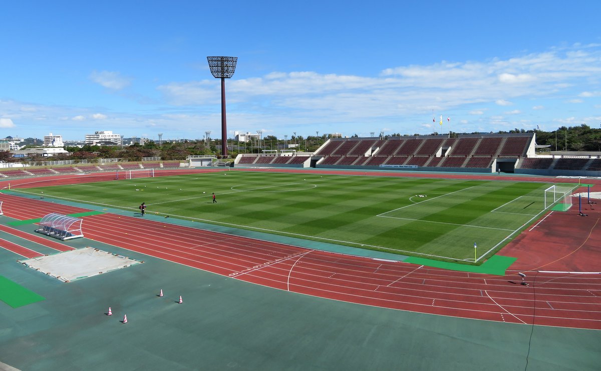 沖縄県総合運動公園陸上競技場的場內:紅色跑道圍著綠色草皮足球場,對面是酒紅色座椅的看台與一支照明塔,晴天藍天