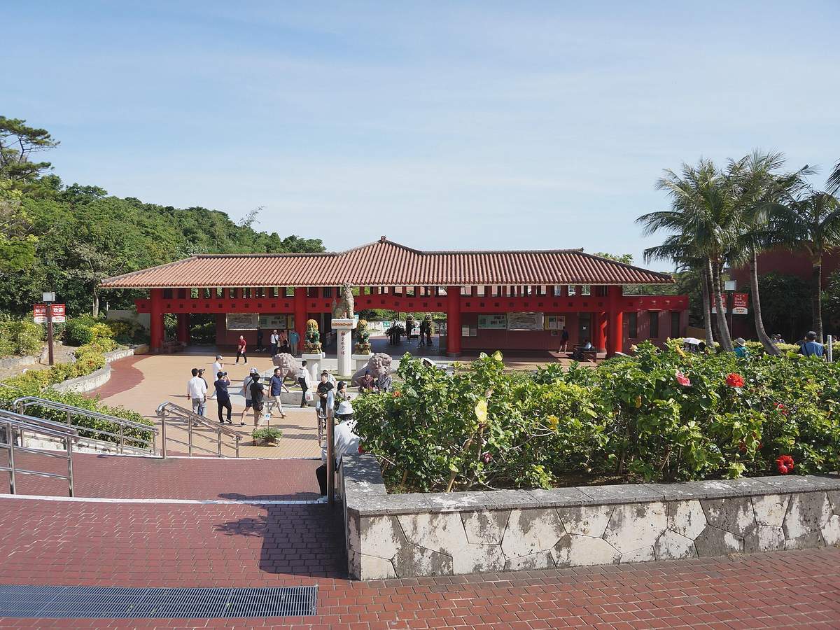 おきなわワールド入口的售票建築：紅色圓柱撐著橫長的紅瓦屋頂，前方廣場立著石獅與一根白色柱子，左側有往下的階梯，右邊是扶桑花叢與椰子樹