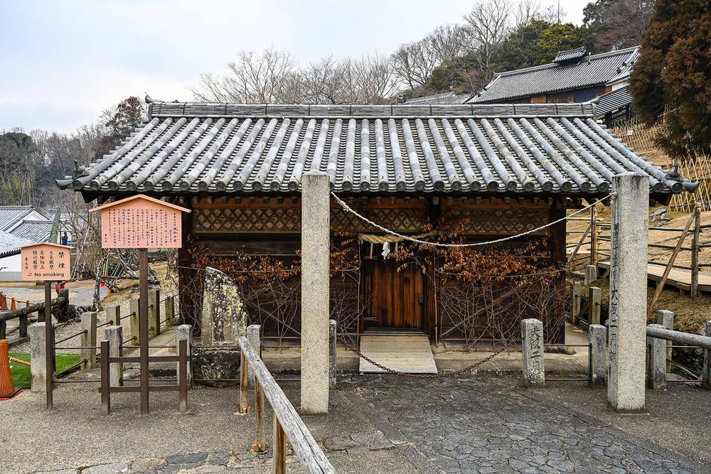 閼伽井屋正面:瓦頂木造小屋,門前拉著粗注連繩,兩根石柱與鐵鍊圍住入口,左邊立著寫有「閼伽井屋」的解說牌與「禁煙 火気厳禁」告示牌,右後方是竹柵圍起的草坡
