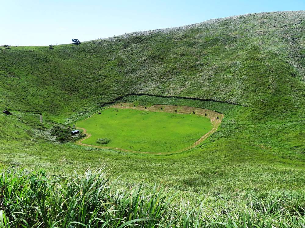 從火山口邊緣往下看大室山火山口:碗底是一塊平坦的鮮綠色草地,邊緣排著一列小小的箭靶,旁邊有一間小屋,四周是長滿草的陡坡