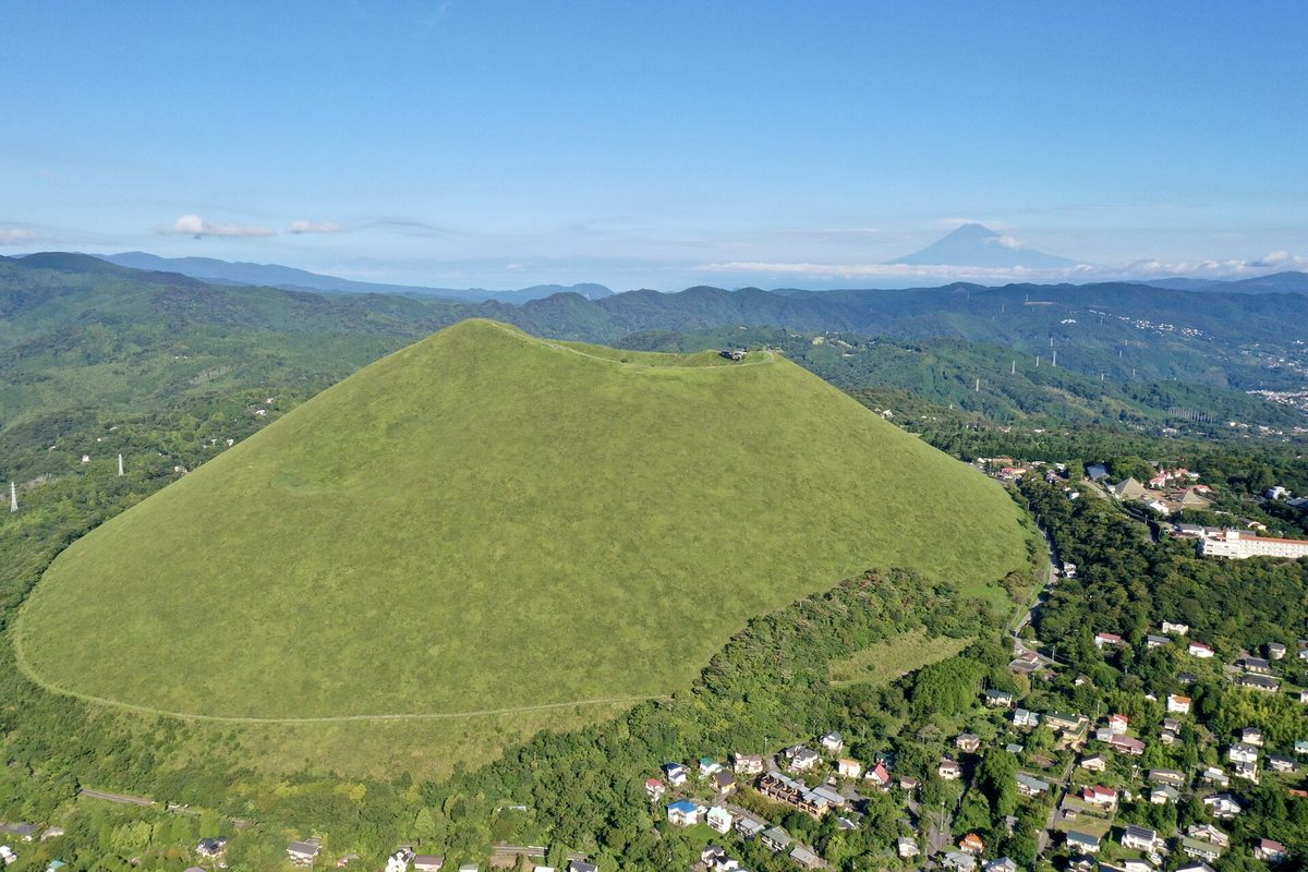 空拍九月的大室山:整座碗形山被翠綠的草覆蓋,山頂邊緣有一棟小建物,山腳繞著一條小路,右下是住宅區,遠方地平線上是富士山