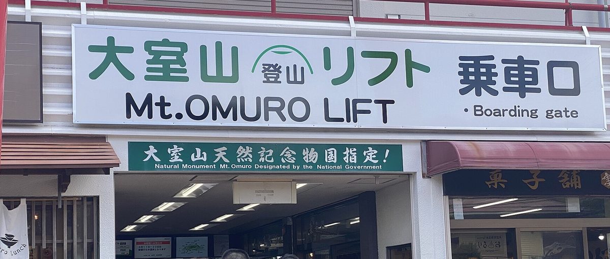 大室山登山吊椅乘車口的白色大招牌,綠字「大室山 登山リフト」與黑字「乗車口 Mt.OMURO LIFT Boarding gate」,下方綠色橫幅寫「大室山天然記念物国指定!」,右邊是一間菓子舖