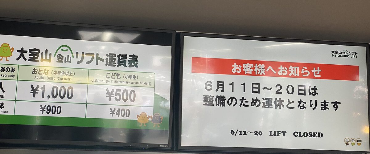 吊椅乘車處天花板下的兩塊螢幕:左邊是「大室山登山リフト運賃表」,大人 1,000 日圓、小學生 500 日圓、團體 900 與 400 日圓;右邊是「お客様へお知らせ 6月11日〜20日は整備のため運休となります」的停駛公告