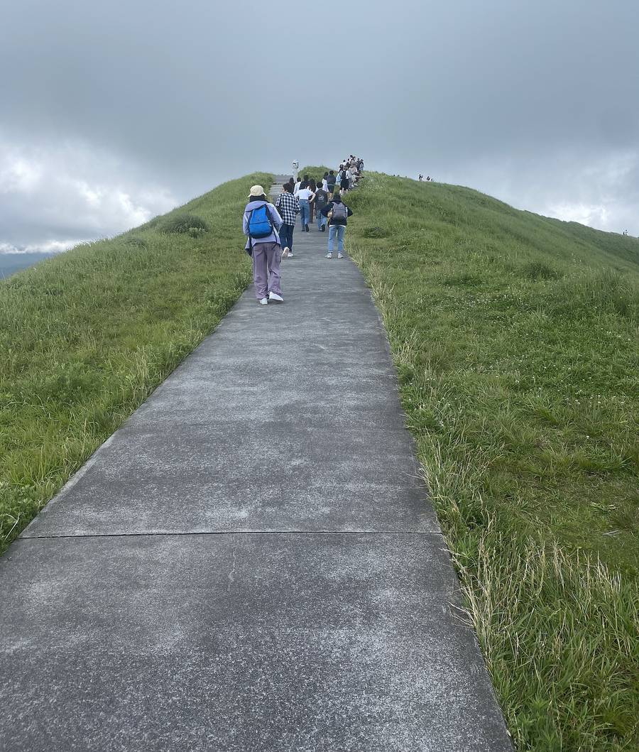 大室山山頂お鉢めぐり步道:一條水泥步道沿著長滿綠草的火山口邊緣往上坡延伸,幾位遊客背對鏡頭走在步道上,天空是灰色的雲