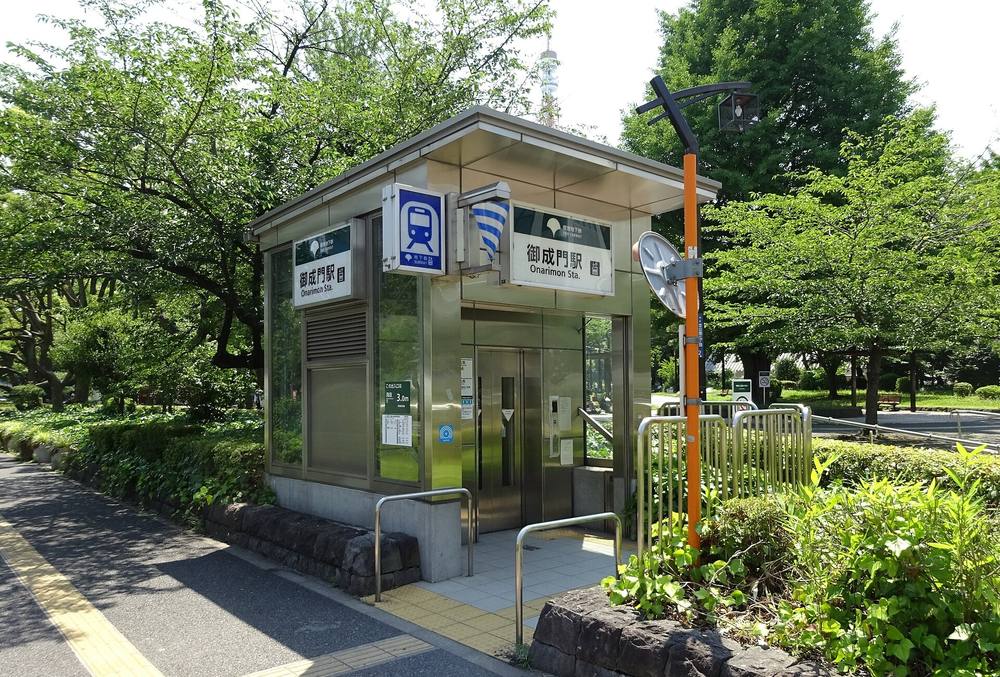 都營三田線御成門站 A6 出入口的電梯棟:人行道旁一座不鏽鋼外牆的小屋,掛著兩塊「御成門駅 Onarimon Sta.」站名牌與藍色地下鐵標誌,四周是公園的綠樹,屋頂後方露出東京鐵塔的塔尖