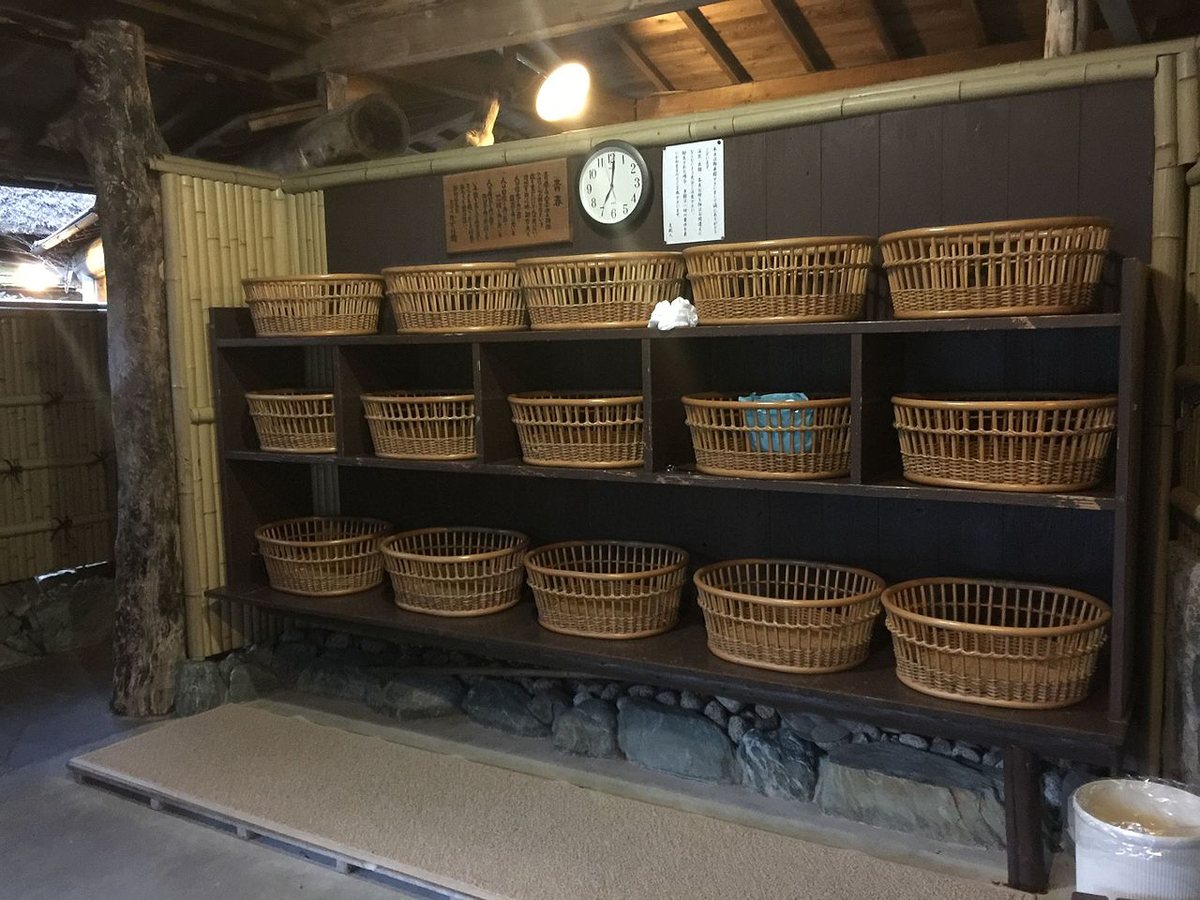 A corner of a Japanese onsen changing room: a dark timber rack of three shelves holding fifteen woven wicker baskets, a white bin at the lower right, and above the rack a plank wall carrying a vertically written wooden plaque, a round wall clock and a vertically written paper notice, with a bamboo screen to the left