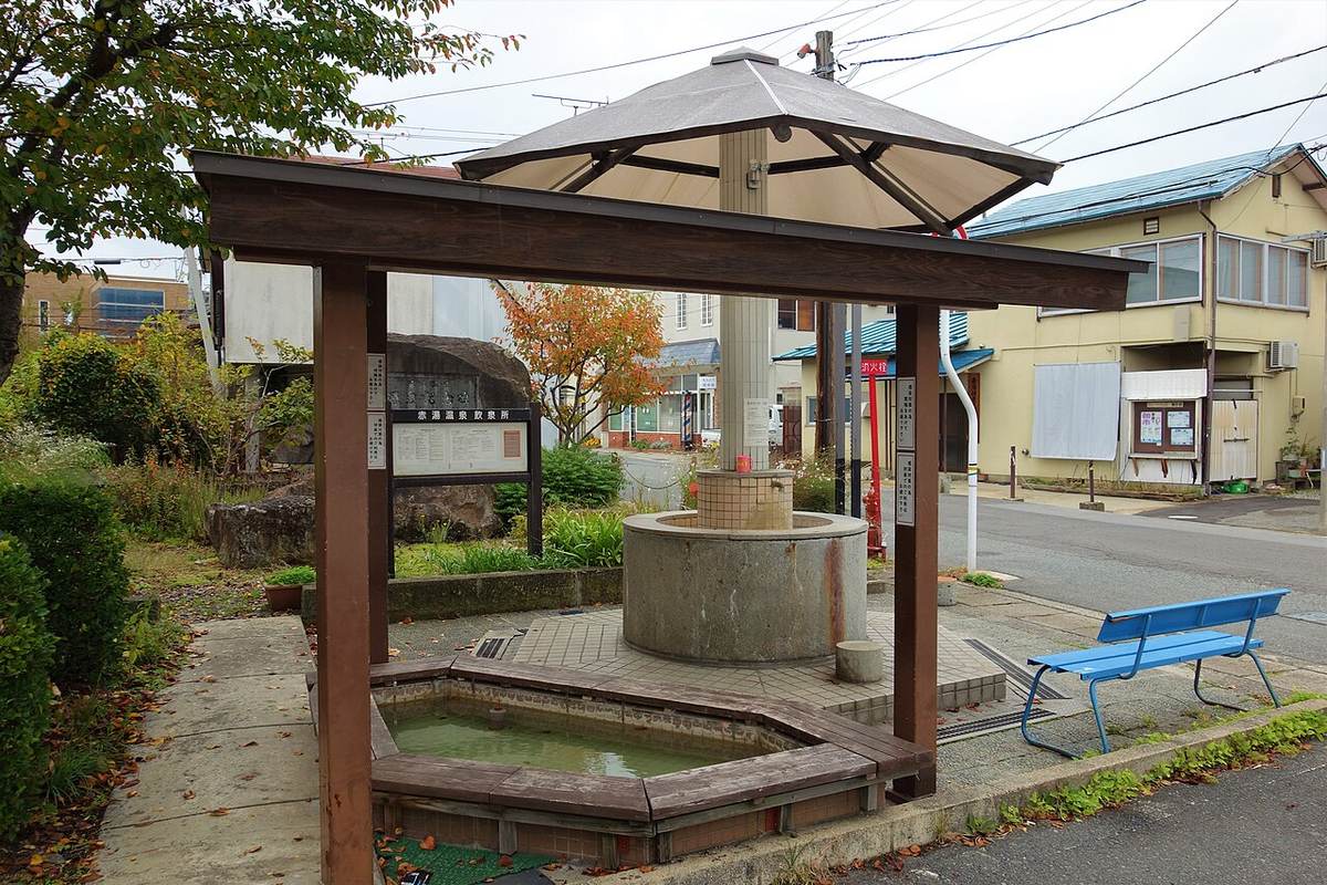 A street-side drinking-water station: a square canopy on timber posts sheltering a cylindrical concrete plinth topped with a tiled spout, with an octagonal timber-framed footbath of pale green water in front. A wooden signboard to the left reads 赤湯温泉 飲泉所 above a dense block of explanatory text, flanked by two narrow vertical signs