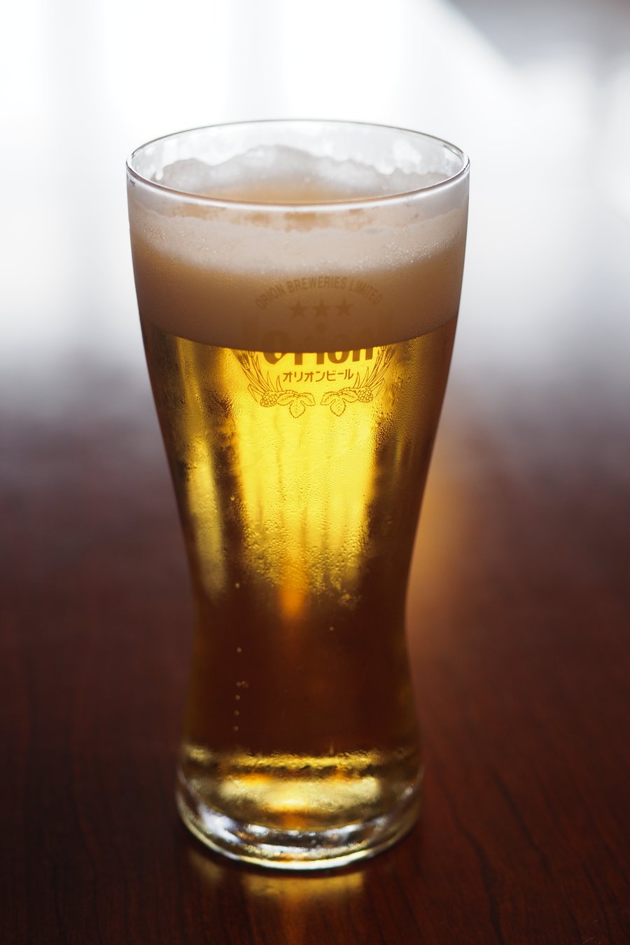 Orion Happy Park 試飲的一杯生啤酒:深色木桌上一只倒滿金黃色啤酒的細腰玻璃杯,頂端一層綿密白色泡沫,杯身印著三顆星、Orion 字樣、「オリオンビール」與啤酒花圖案,背景是明亮的窗光