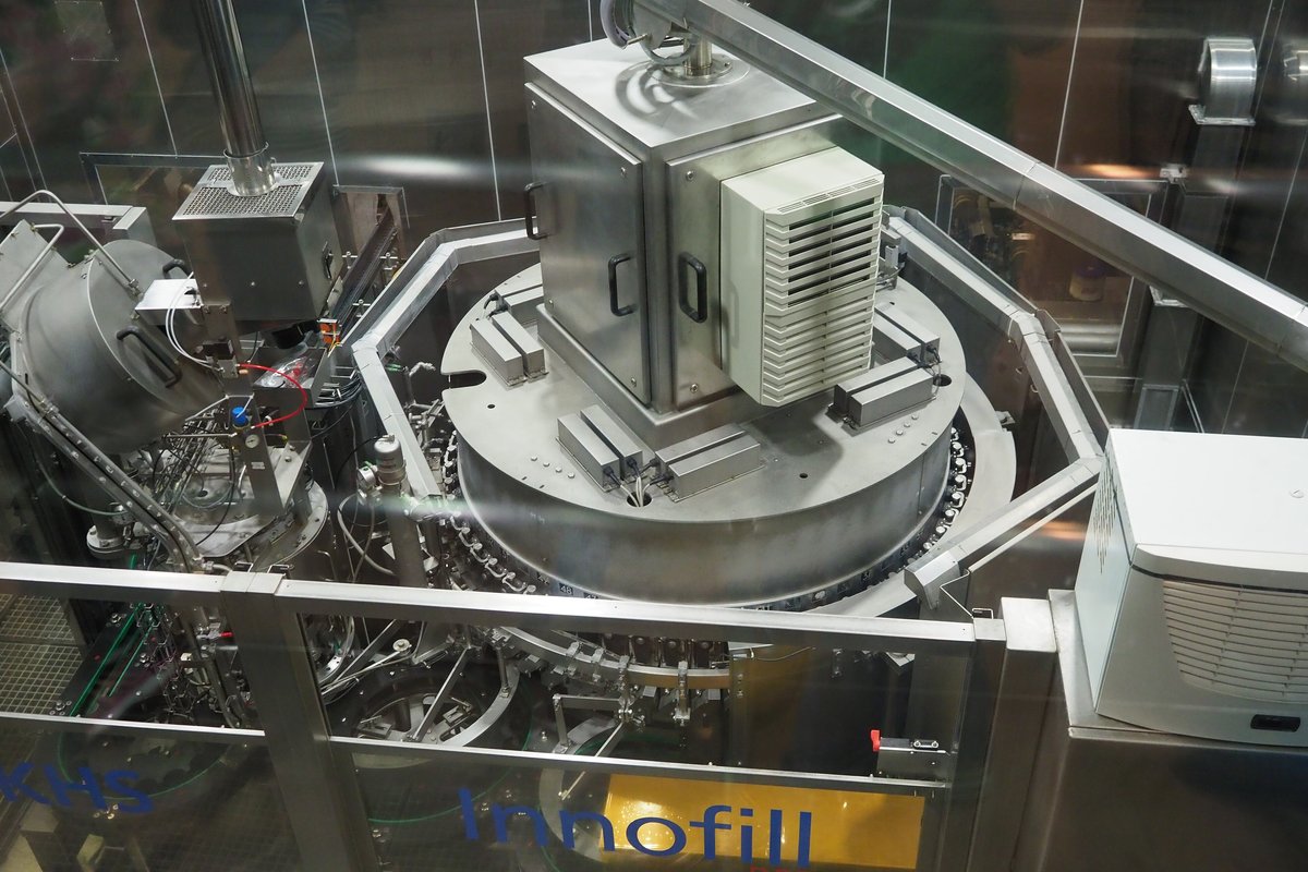 Orion 啤酒名護工廠產線上的一台不鏽鋼充填機,隔著玻璃從斜上方拍:中央是一個大圓盤狀的旋轉台,上面架著一個方形不鏽鋼箱與白色散熱箱,四周是管線、護欄與另一台白色機箱,下方護板印著藍字 KHS 與黃底的 Innofill;畫面裡沒有人