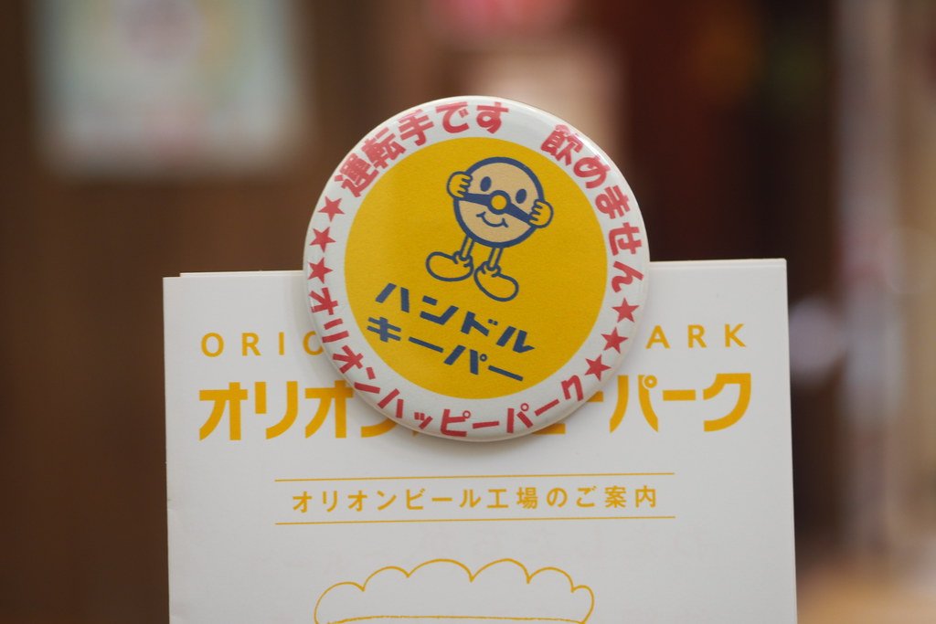 一枚圓形別針徽章夾在 Orion Happy Park 的白色導覽手冊上:黃底中央是一個藍色細線勾邊的圓臉卡通人物與藍字「ハンドルキーパー」,外圈紅字寫「運転手です 飲みません」與「オリオンハッピーパーク」;手冊封面印著橘字「オリオンハッピーパーク」與「オリオンビール工場のご案内」,背景模糊