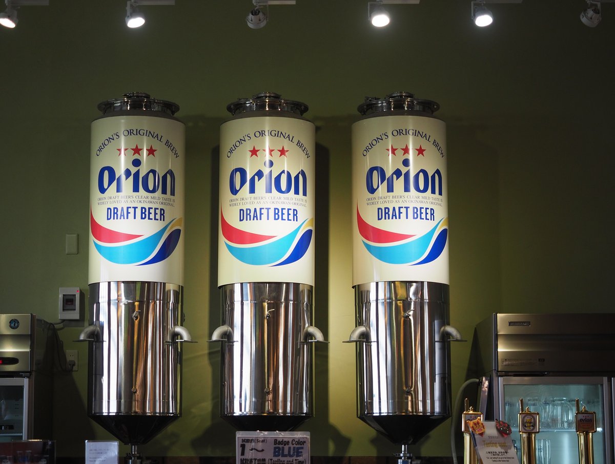 Orion Happy Park 試飲會場的吧台:橄欖綠的牆前並排三座下半部是不鏽鋼、上半部包著米白色外殼的圓柱形啤酒槽,外殼印著 ORION'S ORIGINAL BREW、三顆紅星、藍字 Orion 與 DRAFT BEER;吧台上立著寫「試飲席(Seat) 1〜 Badge Color BLUE」的小牌,右邊是一排金色啤酒拉把、玻璃杯冰櫃,以及寫著「Tasting seat 工場見学後、試飲会場へご案内します」的立牌