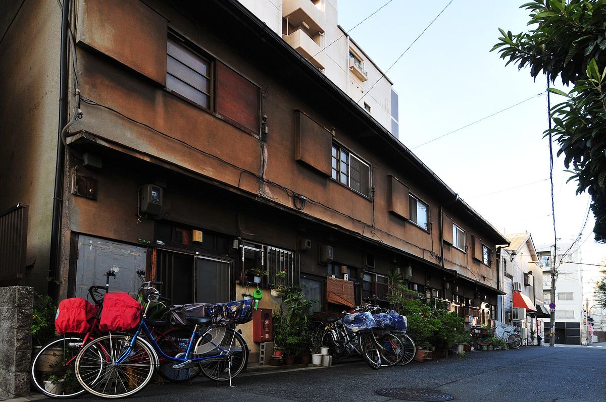 A residential back street near Tenmabashi in Osaka: a long two-storey row of older bunka-jutaku flats, bicycles under rain covers and rows of potted plants along the frontage, a new high-rise block behind