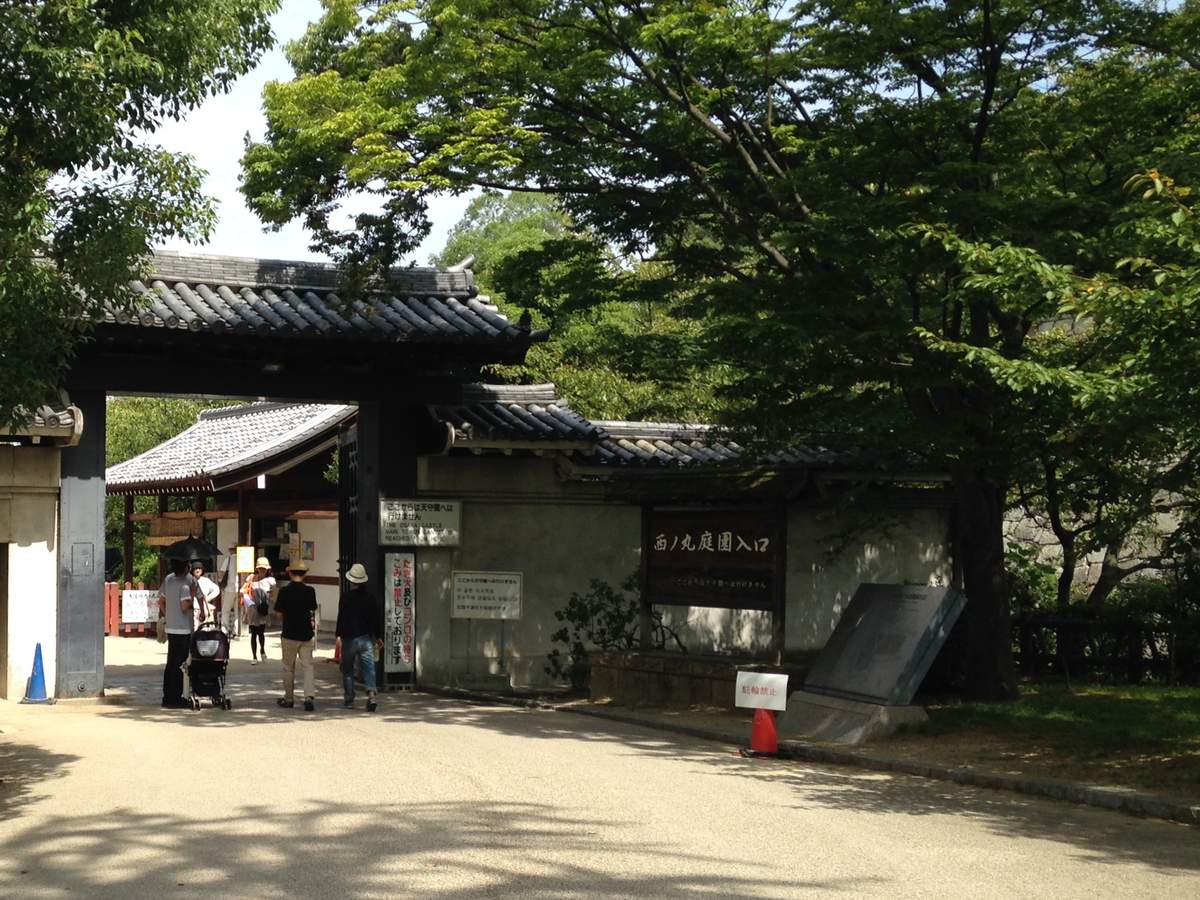 白天的西之丸庭園入口:瓦頂木門,右邊黑色木牌寫「西ノ丸庭園入口」,門柱旁白牌寫從這裡到不了天守閣,遊客背對鏡頭往裡走
