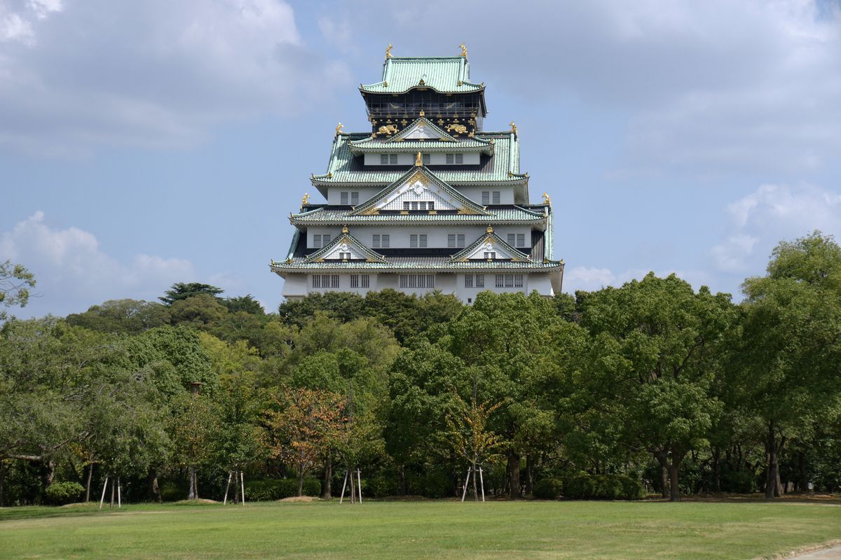 白天的大阪城西之丸庭園:前景一大片草坪,後面一排樹林,天守閣從樹林上方露出來,天空多雲