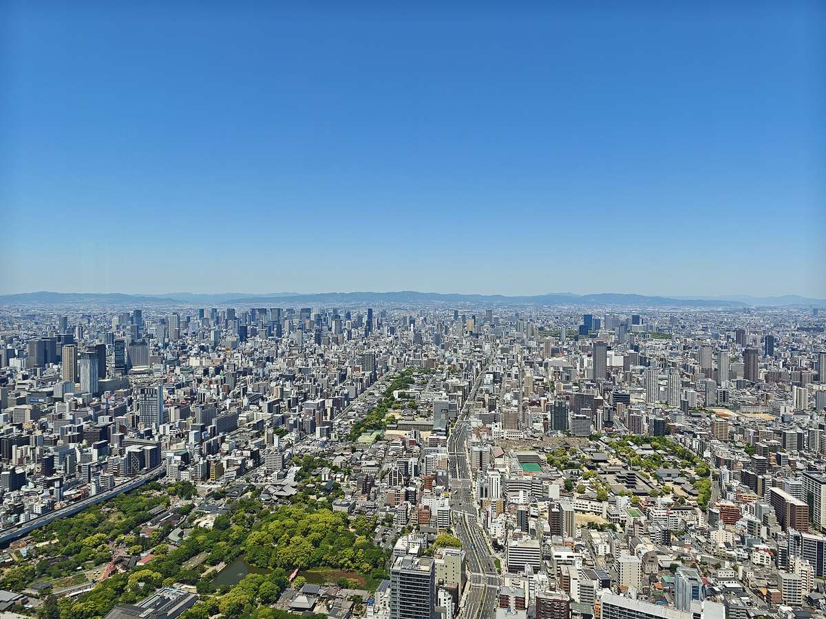 Osaka seen from high up in Abeno Harukas: low-rise blocks and towers run all the way to the horizon, a line of distant mountain ridges is sharply outlined, the sky is cloudless deep blue, and a park with a pond fills the lower left