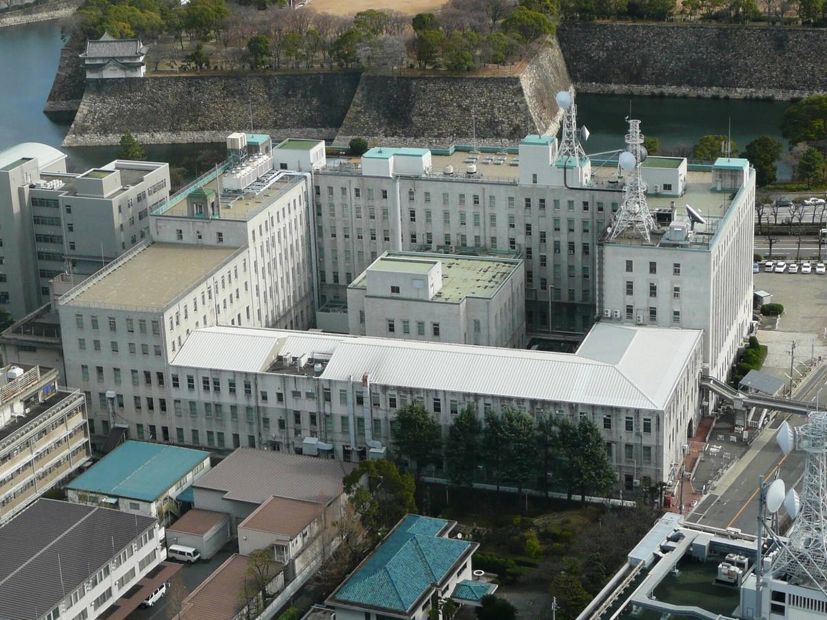 從高處俯瞰大阪府庁本館:一棟灰白色、口字形的老式辦公大樓,屋頂有通訊鐵塔;大樓後方隔著一條路就是大阪城的護城河與高石垣,石垣上有一座白色櫓樓