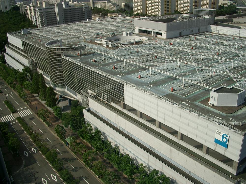 從高樓俯瞰インテックス大阪 6 號館:方正的大型展館,屋頂是停車場與白色鋼架,外牆角落有藍底白字的「INTEX OSAKA 6」標示,館旁是有行道樹的寬馬路