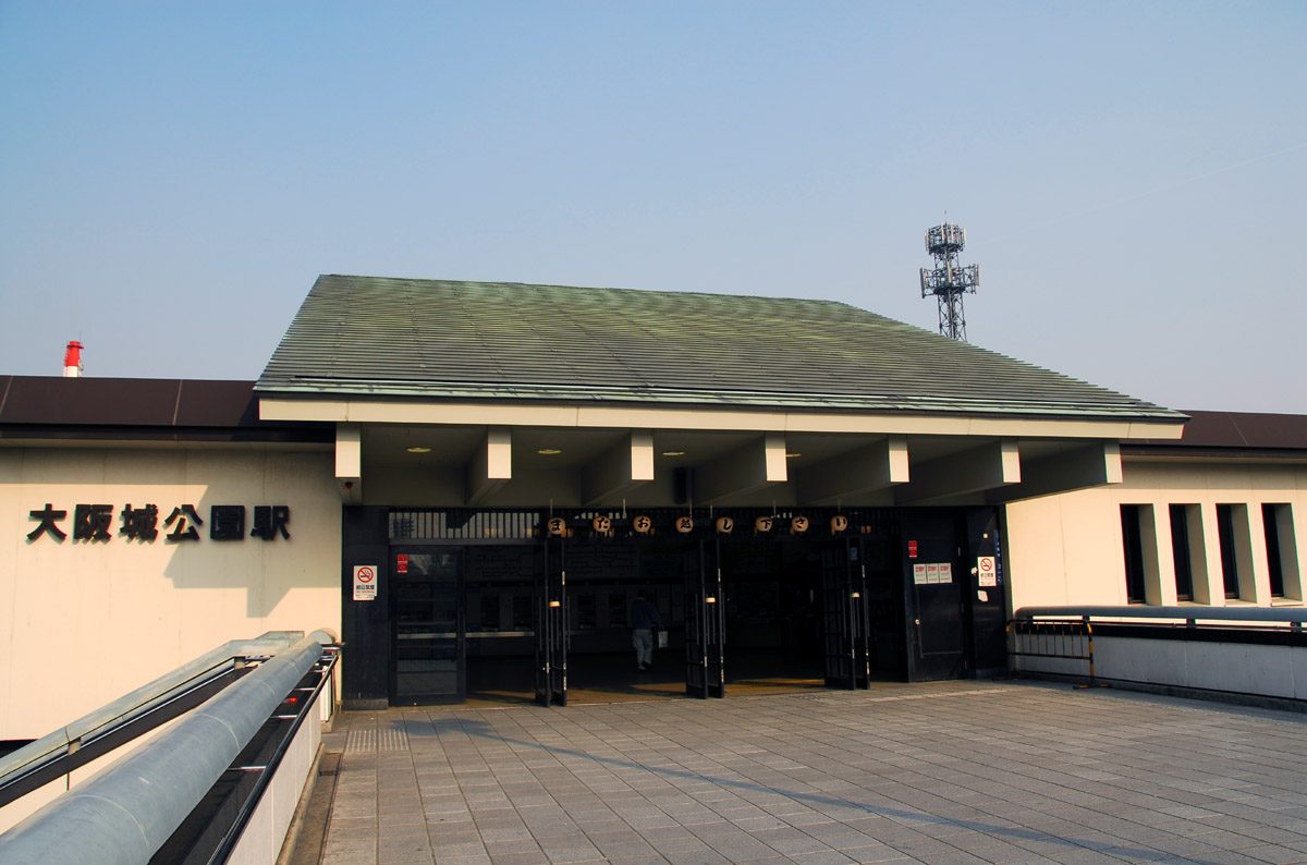 JR 大阪城公園站的站房:深綠色的大斜屋頂,左邊白牆上寫著「大阪城公園駅」,正面是一排黑色格柵門,前面是鋪石板的站前通道,晴天