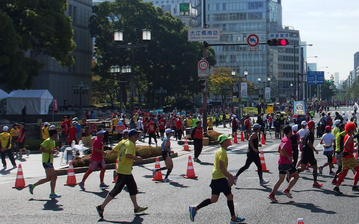 大阪馬拉松跑在御堂筋上:大江橋南詰路口,跑者成群往畫面右後方跑,路中間排著三角錐,左側人行道邊有桌子與穿背心的工作人員,後方是大阪市役所旁的綠樹與高樓,路牌寫著「大江橋南詰」