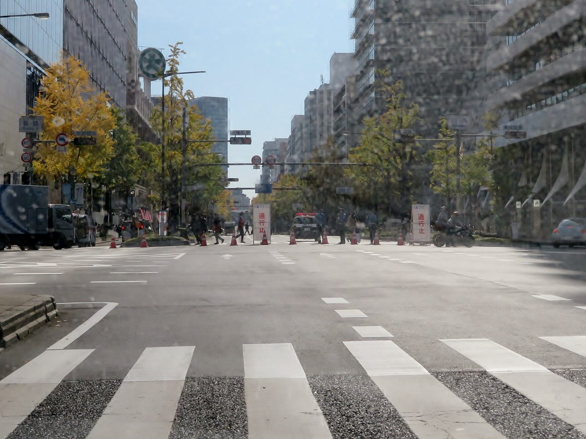 比賽日的御堂筋新橋路口:寬闊的車道空無一車,遠處一排三角錐與兩塊寫著「大阪マラソン 通行止」的白色看板把路擋住,看板中間停著一輛警車,兩側是銀杏行道樹與商辦大樓;照片隔著有水痕的車窗拍