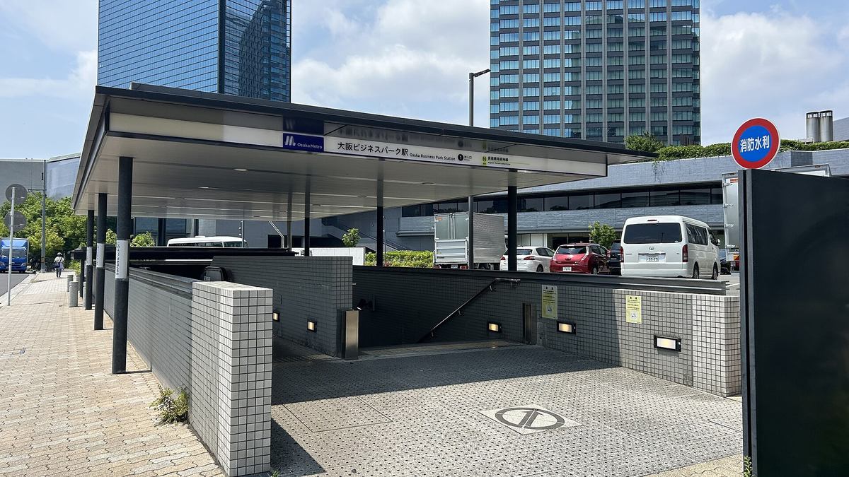 Osaka Metro 大阪ビジネスパーク站的 1 號出入口:平頂雨棚下是往地下的階梯,棚簷寫著站名與「1 出入口」、長堀鶴見緑地線,後方是兩棟玻璃帷幕高樓