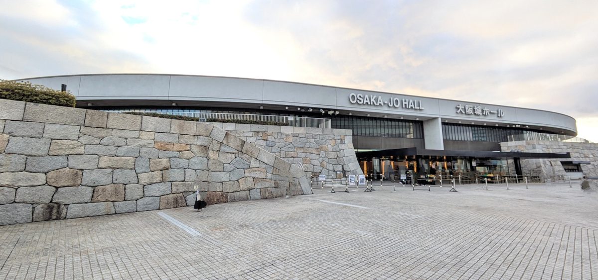 大阪城ホール正面:弧形的白色屋簷上寫著「OSAKA-JO HALL」與「大阪城ホール」,左側是大石塊砌的石牆,前面是空曠的石板廣場,傍晚多雲