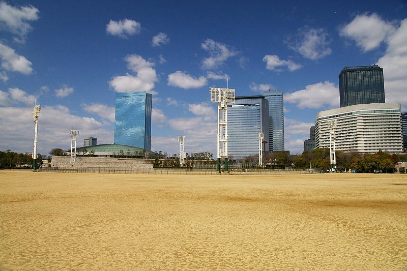 大阪城公園的太陽の広場:一整片黃土空地,周圍立著幾支照明燈柱,左後方是綠色圓頂的大阪城ホール,右後方是大阪商務園區的玻璃帷幕高樓與一棟白色飯店,藍天白雲