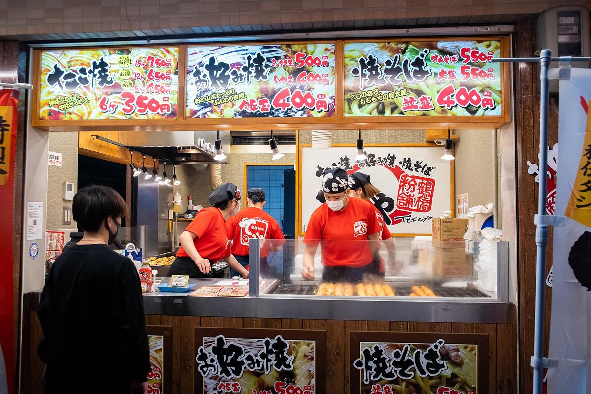 Street-food stall in Osaka's Shinsekai district: staff in red shirts griddling takoyaki behind the counter under a menu board for takoyaki, okonomiyaki and yakisoba