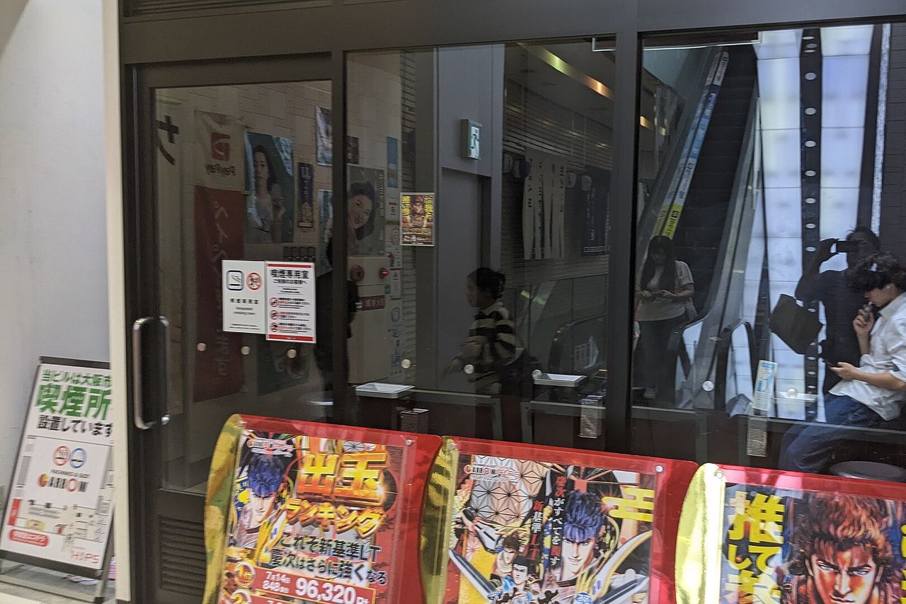 A glass-walled indoor smoking room in the Namba Hips building, with a legal notice and usage rules on the door and a standing board saying the building provides a smoking area