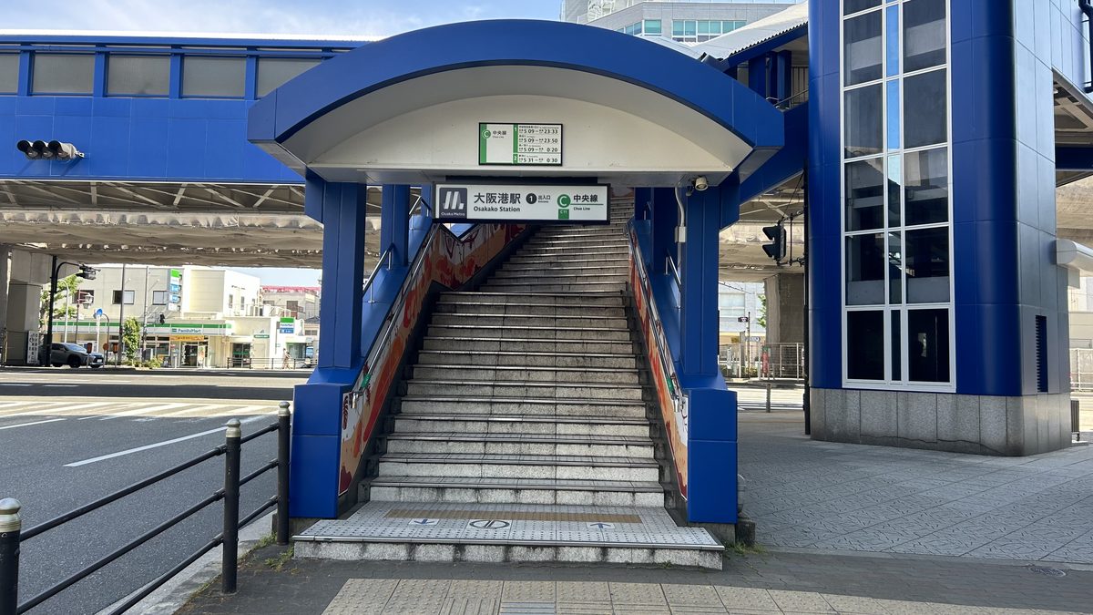 大阪港站 1 號出入口,藍色拱形雨棚下是一段往上通往高架車站的長樓梯,門楣上的站牌寫「大阪港駅 1 出入口 中央線」,旁邊是藍色的高架車站建築