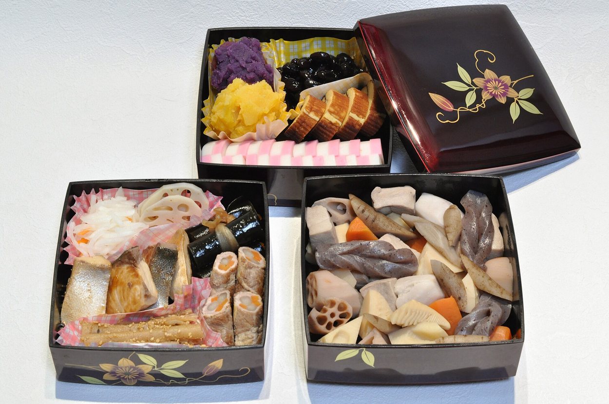 Osechi New Year food arranged in stacked lacquer jubako boxes: black soybeans, sweet chestnut paste, rolled omelette, pink and white fish cake, simmered vegetables and grilled fish