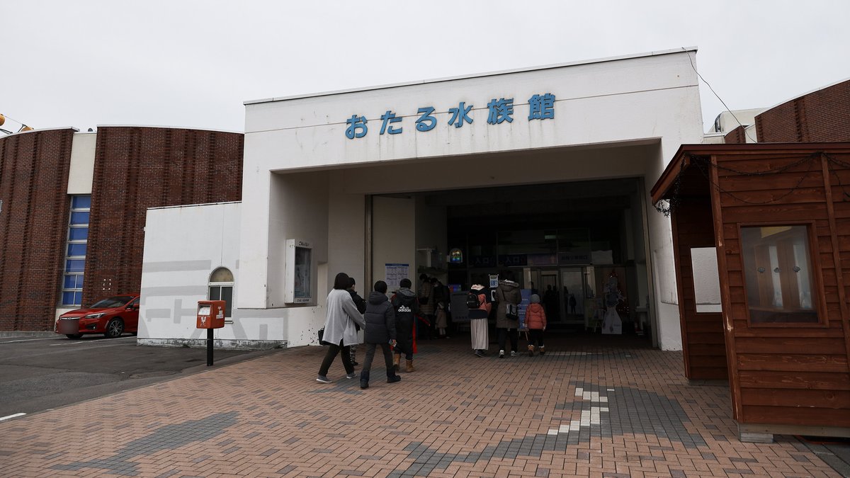 おたる水族館本館入口：白色門廊上方是藍色「おたる水族館」字樣，兩側紅磚圓弧外牆，遊客穿冬衣走進入口