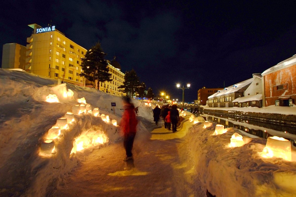 The Otaru Canal venue of Yuki Akari no Michi: snow candles line both sides of a trodden path as visitors walk between them, with snow-capped stone warehouses to the right