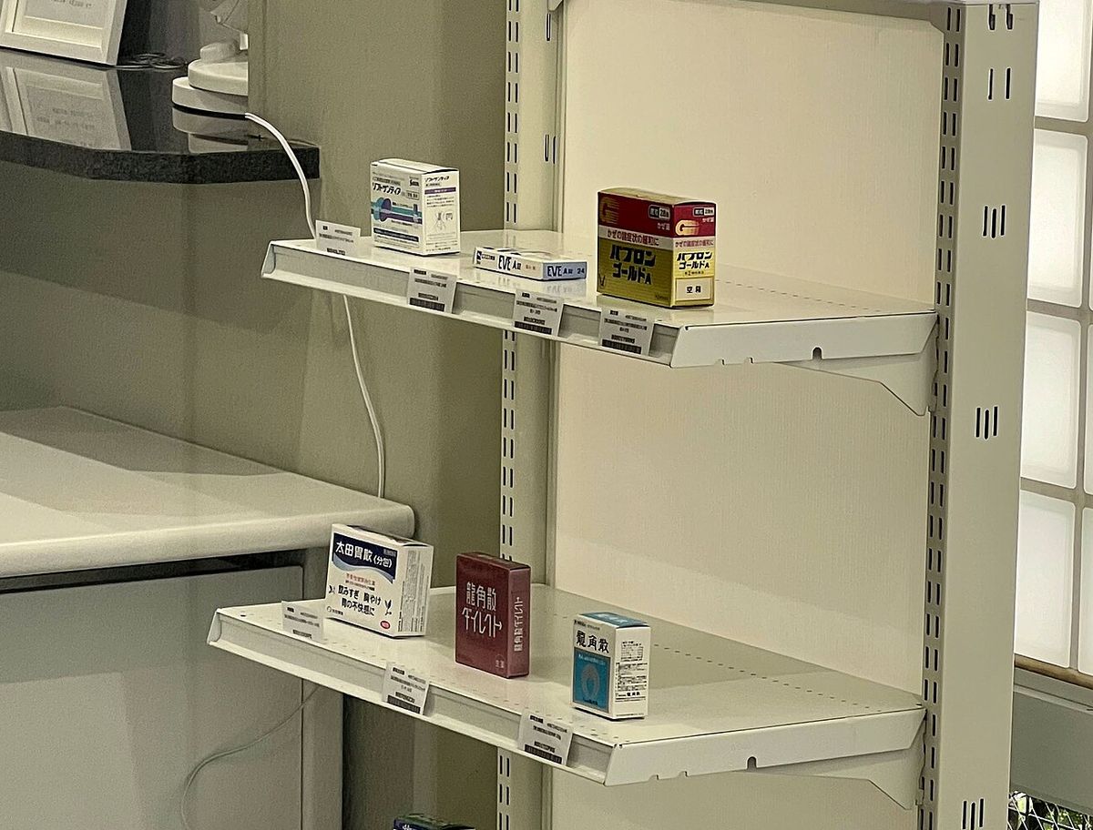 White metal shelving in a small Japanese pharmacy holding only three boxed medicines on the upper shelf; the cold-remedy box on the right carries a white sticker reading kūbako, empty box, and each carton has a small printed card with its name and barcode slotted into the shelf edge in front of it. Three more boxes sit on the middle shelf and the bottom shelf is bare.