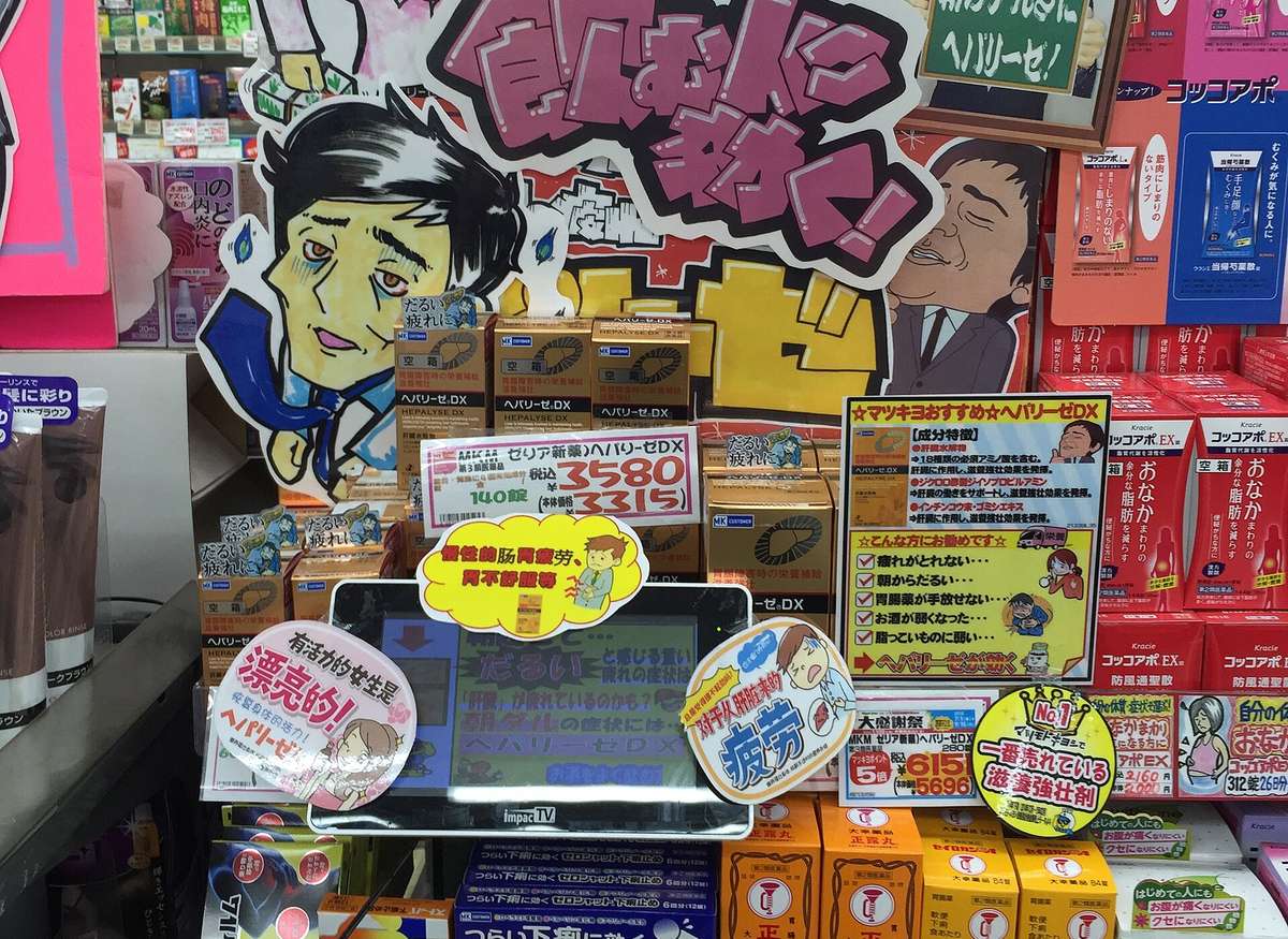 The OTC medicine section of a Japanese drugstore: shelves packed with boxed tonics, stomach remedies and anti-diarrhoeals, hand-drawn recommendation boards and red handwritten price cards propped between them, and several display boxes carrying a white sticker reading kūbako, empty box.