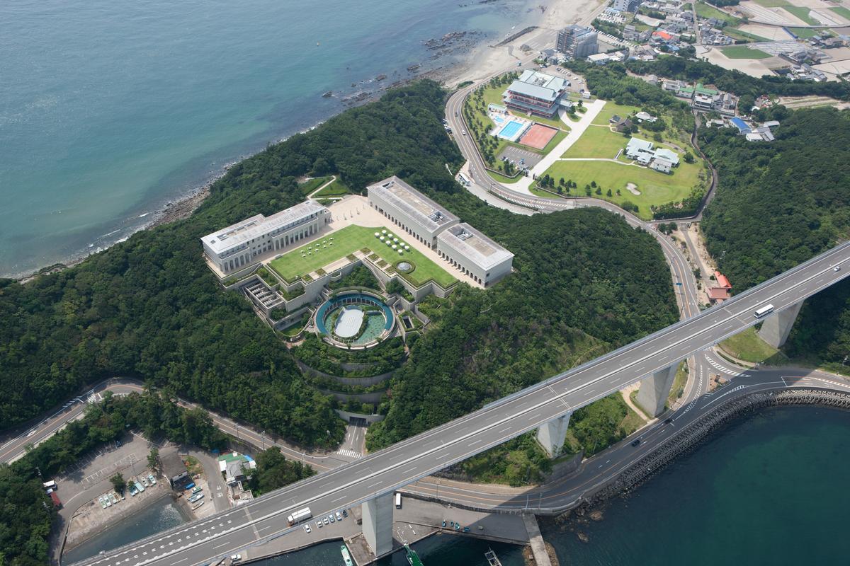 從空中往下看鳴門的海岸:森林小山頭上是美術館的白色建築群與方形草坪,往下一層是圓形的戶外展區,山腳是正門,右下方一條高架高速公路沿海岸延伸過去