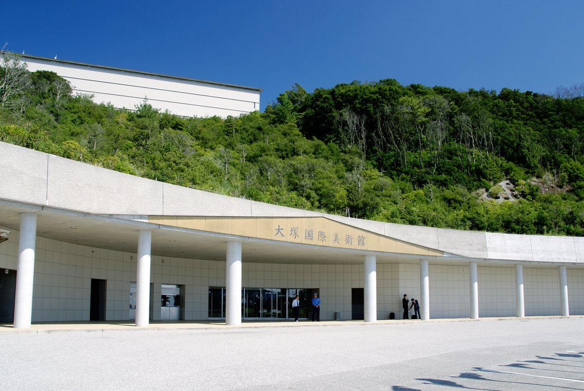 大塚國際美術館正門:山坡下一排白色圓柱撐著的弧形長屋簷,屋簷正中寫著「大塚国際美術館」,玻璃門口站著幾個人,背後山頂上露出美術館的白色建築
