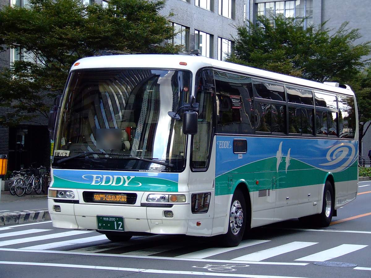 一輛白底、藍綠色波浪塗裝的徳島巴士高速巴士,車頭寫著「EDDY」,擋風玻璃下的跑馬燈顯示「鳴門・徳島駅前」,行駛在大阪市區的斑馬線上