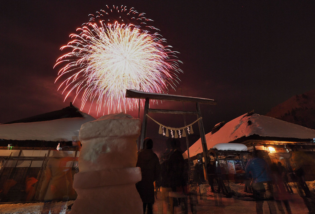 大內宿雪祭的煙火：紅白色的煙火在夜空炸開，前景是一座雪堆成的燈籠與掛著注連繩的木鳥居，兩側是積雪的茅葺屋頂，鳥居下的人群只看得到背影