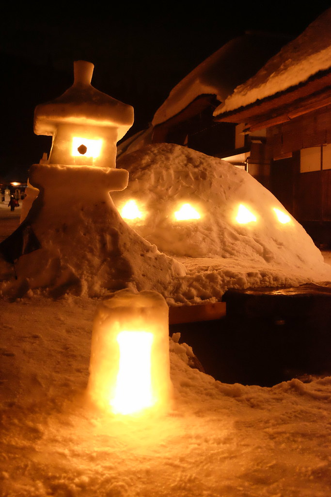 點亮的雪燈籠近照：雪堆成的燈籠與後方雪丘上的四個小洞都透出燭光，最前面一個小雪燈，右側是積雪的茅葺屋簷