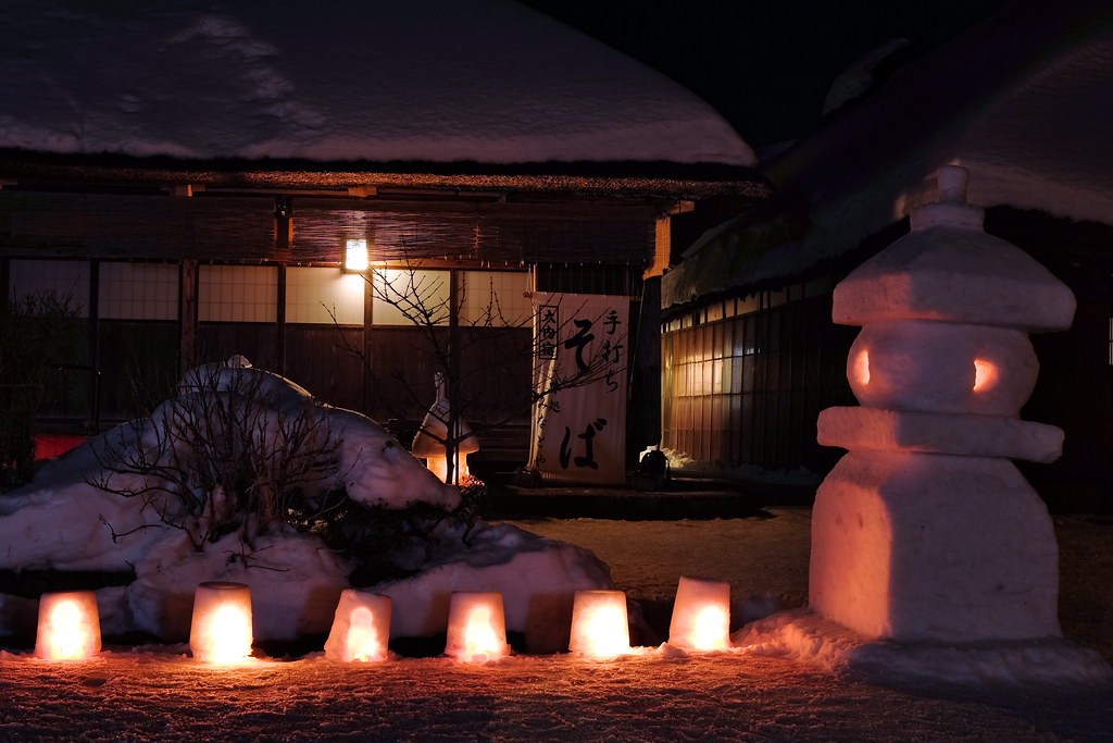 夜晚的大內宿雪祭：屋頂積雪的茅葺老屋前，雪地上一排六個點著燭火的小雪燈，右側一座雪堆成的大燈籠透出橘光，門口掛著「手打ちそば」的布簾