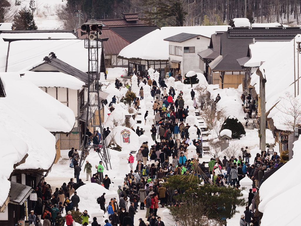 從高處看雪祭白天的大內宿老街：街道中央一整排雪堆成的燈籠，兩側擠滿遊客，左側一座掛著擴音喇叭的鐵塔，屋頂都蓋著厚雪