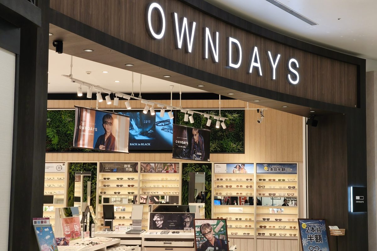 The front of an OWNDAYS store inside a Japanese shopping mall, its lit sign above wall-to-wall frame shelving and a central display table