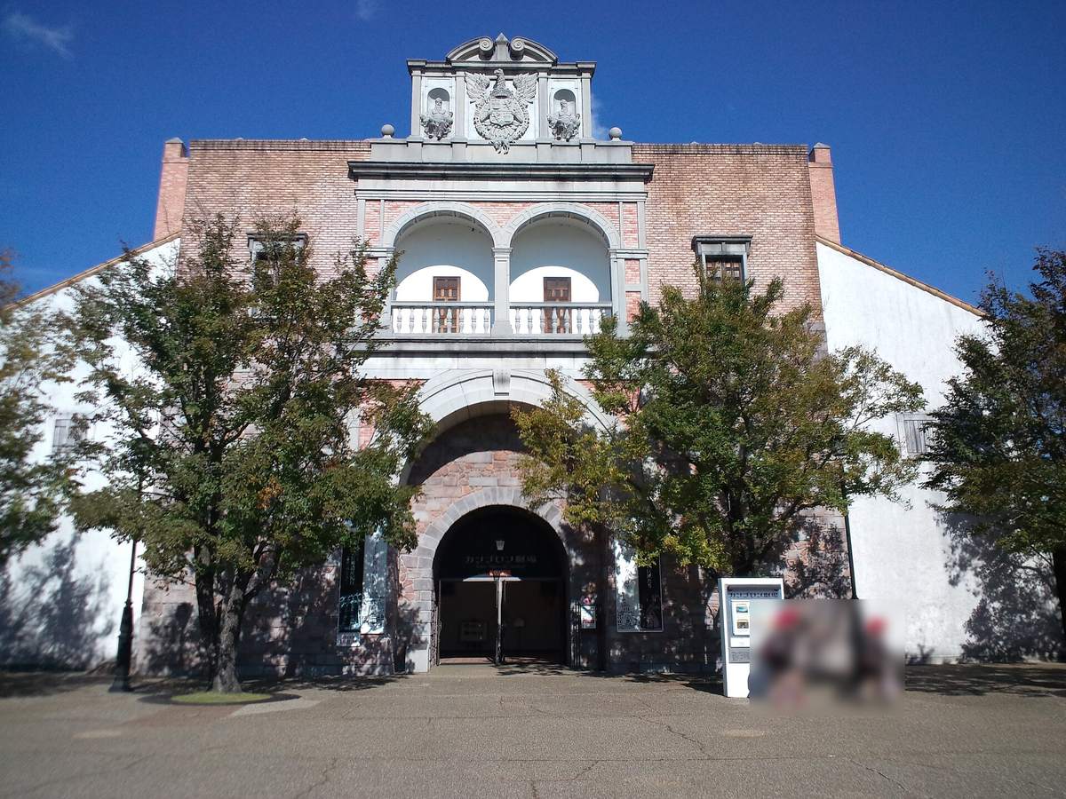 志摩西班牙村的カンブロン劇場：紅磚正面頂上有雙頭鷹紋章，中段是兩個白色拱形陽台，下方是石砌的大拱門入口，兩側種著樹，入口右邊立著劇場的看板