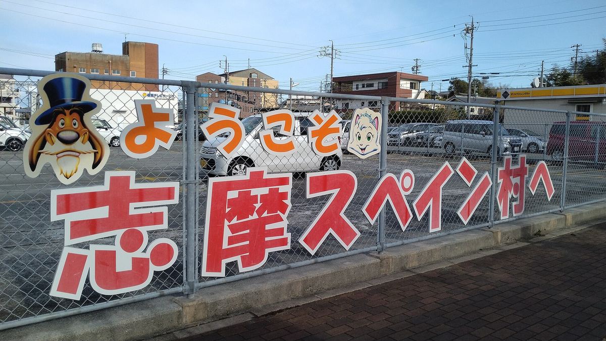 鵜方站前人行道旁的鐵絲網圍欄上，掛著一個個大字拼成的「ようこそ志摩スペイン村へ」紅橘色看板，左右兩端是樂園角色的臉，圍欄後方是停車場