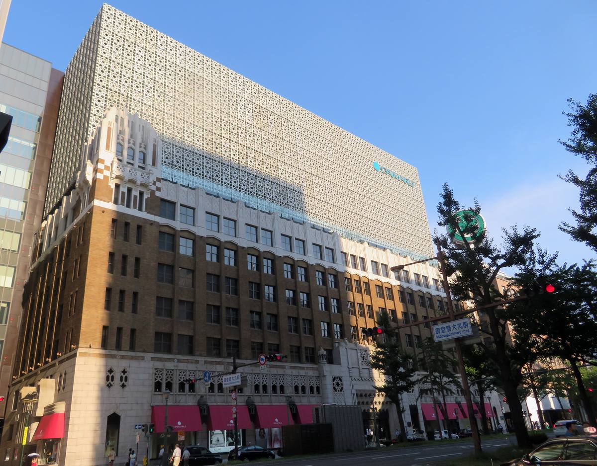 從御堂筋看大丸心齋橋店本館:下半是褐色磚牆與米色石材的舊外牆,一樓整排紅色遮陽棚,上半是白色鏤空花紋的新外牆,右上有綠色的大丸標誌,路口號誌牌寫著御堂筋大丸前